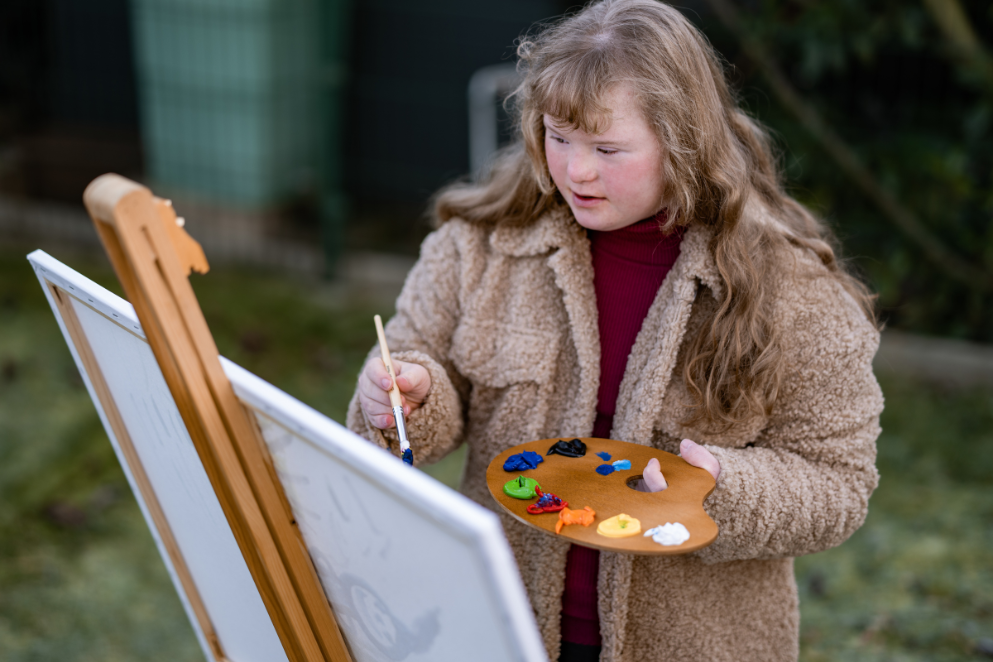 Dziewczynka z paletką i pędzlem w dłoni malująca obraz na płótnie ustawionym na sztaludze