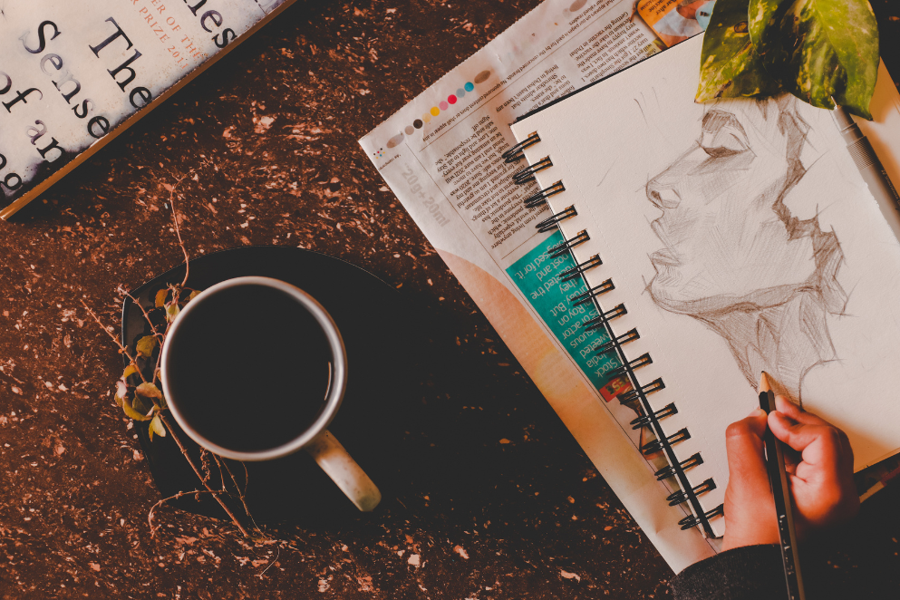 Kobieta używająca szkicownika lezącego na stoliku, na którym znajduje się kubek z kawą i gazety