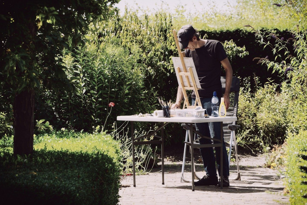 Mężczyzna stojący przy stoliku rozłożonym w plenerze na którym znajduje się sztaluga drewniana z płótnem